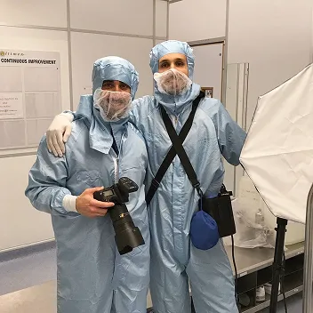 Fede con una cámara, con traje y cofia de sala limpia, abrazando a un colega